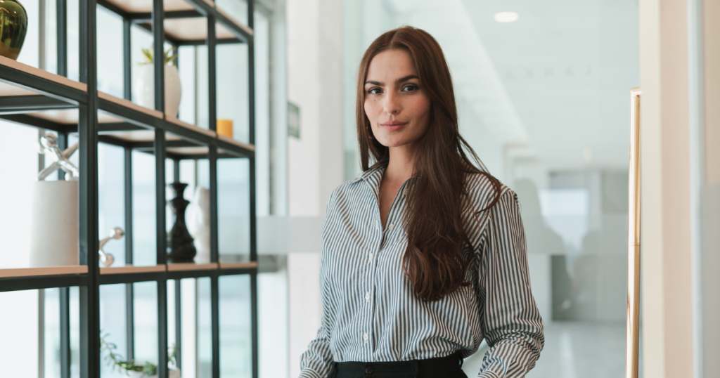 Professional woman with long brown hair in a blue-striped blouse stands by a shelving unit in a bright, modern office.