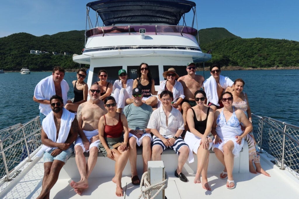 Group of people in swimwear posing for a group photo on the bow of a white boat, with turquoise water and green hills in the background.