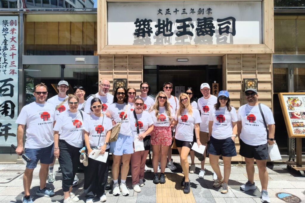 Group of people in white Tokyo 2025 shirts posing together outside a Japanese restaurant with wooden framing and signage.
