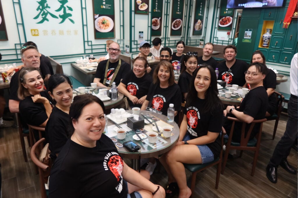 Group of coworkers wearing black shirts with a red logo, seated around a table at an Asian restaurant, smiling at the camera.