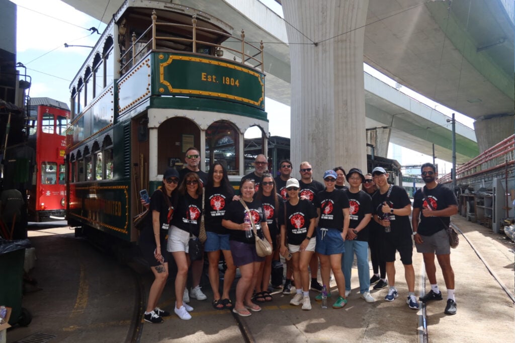 Group of tourists in black shirts posing in front of a vintage green tram car labeled Est. 1904, under elevated highways.