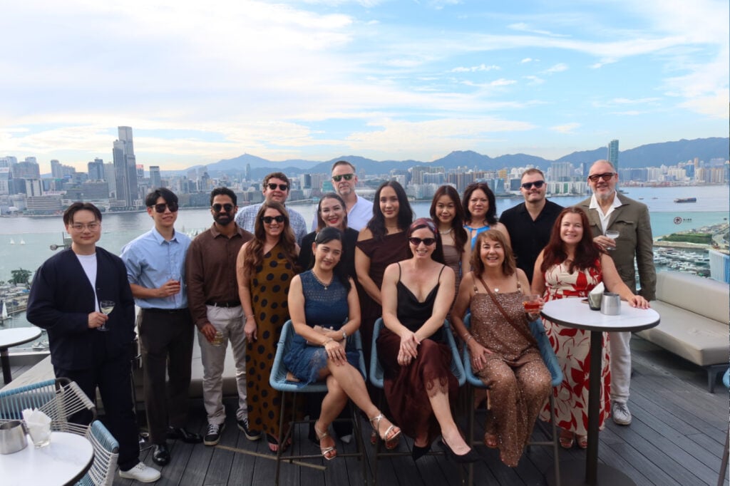 Group of 15 adults posing on a rooftop terrace with a city skyline and water in the background, smiling at the camera during a social event.