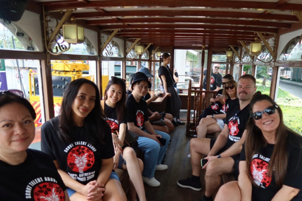 Group of people wearing matching black t-shirts with a red circular logo riding inside a vintage wooden tram, posing for a photo.