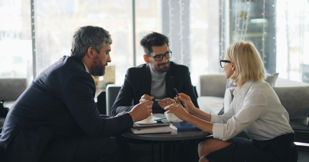 Three professionals in business attire sit around a round table, discussing documents and coffee in a bright, glass-walled lounge area.