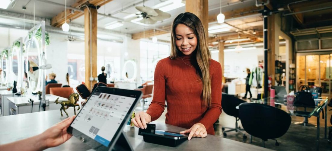 Woman in a rust sweater smiling as she taps a card on a card reader at a checkout counter in a bright, industrial-style shop.